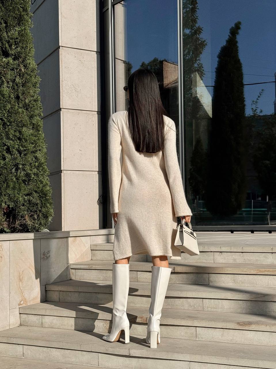 Woman in a white dress and knee-high boots standing on steps outside a building with large windows.
