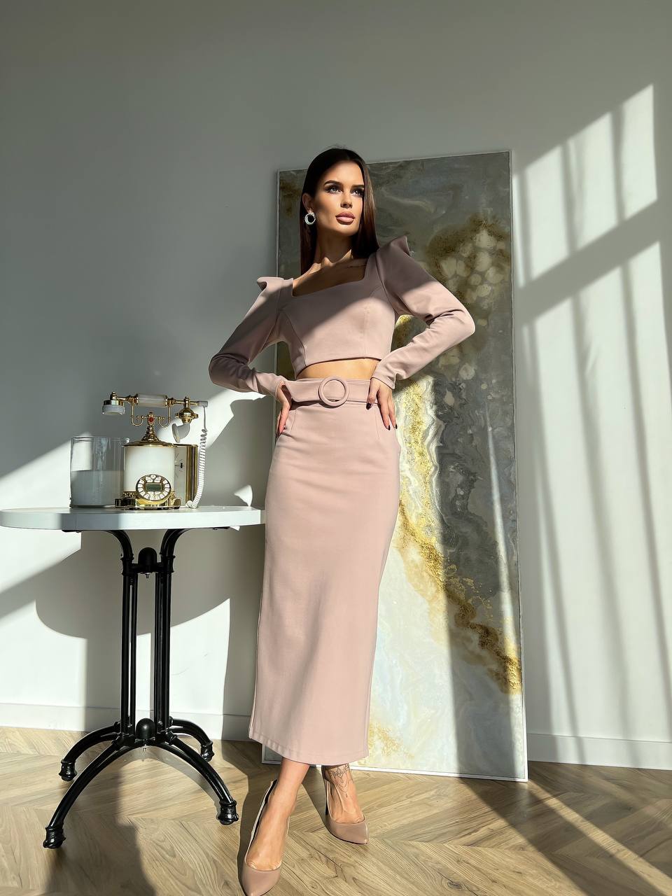 Woman in a stylish outfit standing in a room with sunlight streaming through a window.
