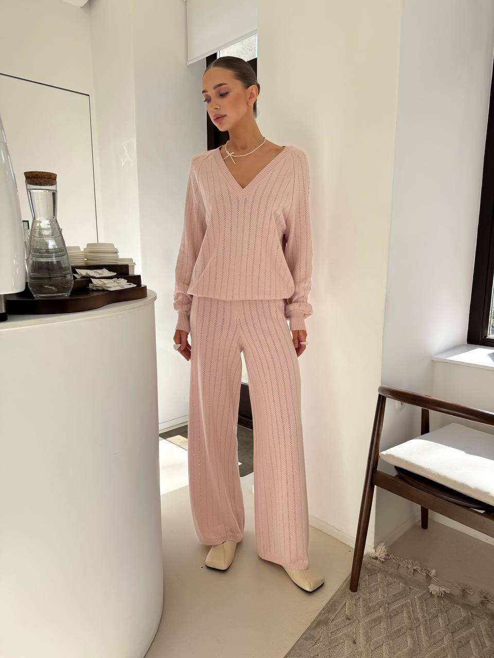 Woman wearing a pink ribbed sweater and pants in a minimalistic room.