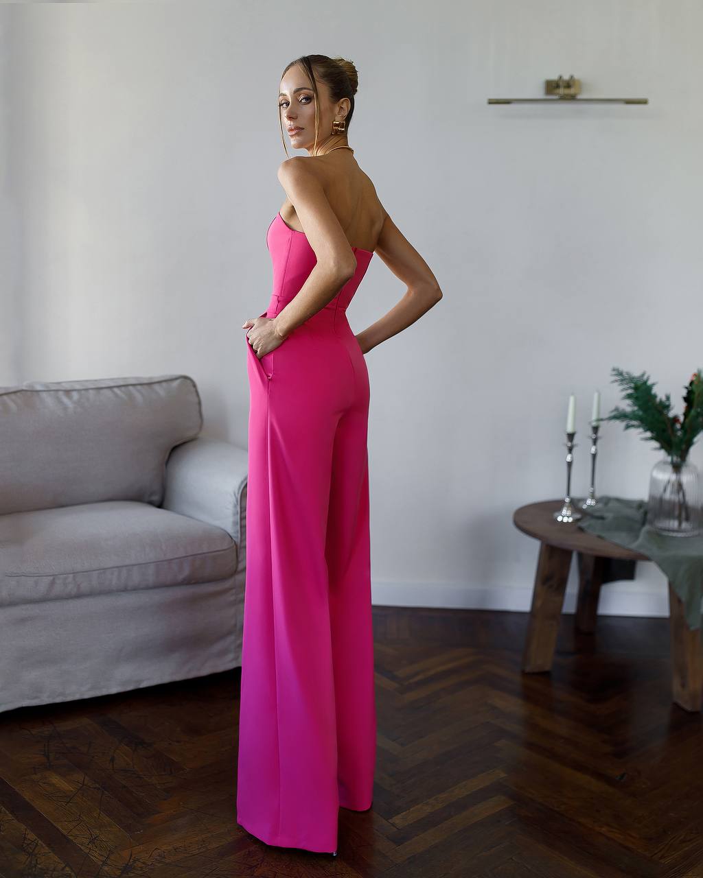 Woman in a bright pink strapless jumpsuit standing in a room with a couch and table.