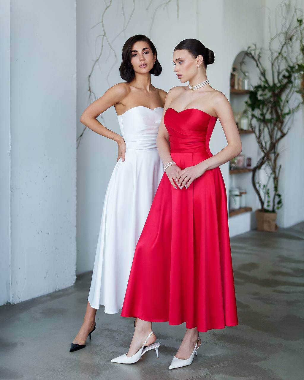 Two women wearing strapless dresses, one white and one red, standing side by side in a room with a plant and shelves in the background.