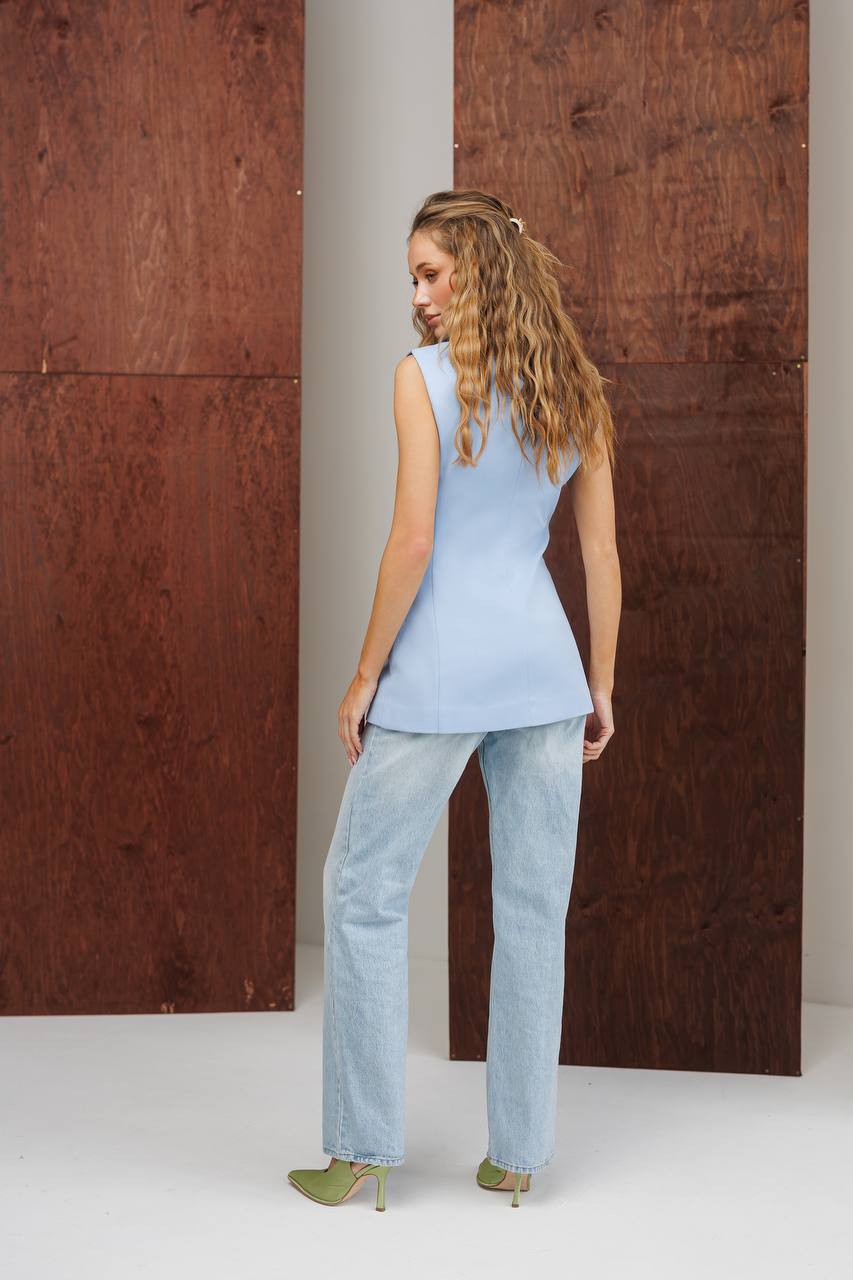 Woman wearing a light blue sleeveless top and jeans standing against a wooden wall.