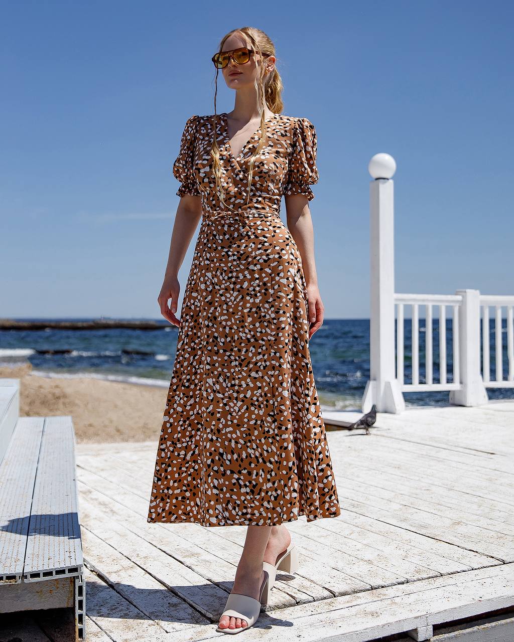 Woman in a floral dress standing on a wooden deck by the ocean.