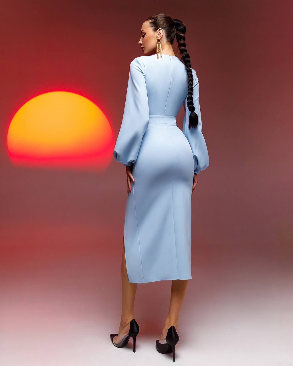 Woman in a light blue dress standing with her back to the camera, facing a sunset.