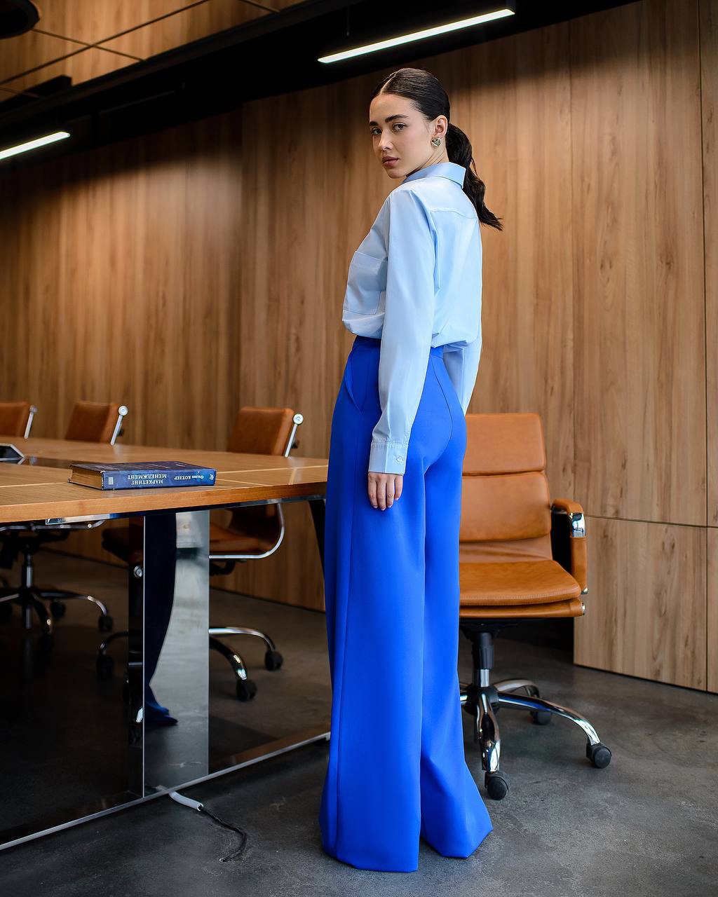 Woman in a light blue shirt and bright blue pants standing in an office setting.