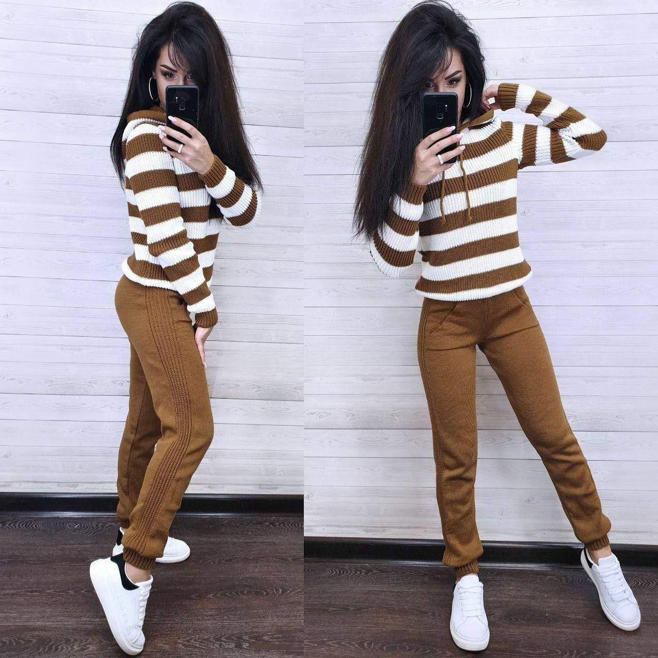 Woman wearing a brown and white striped sweater and brown pants, taking a selfie against a white wall.