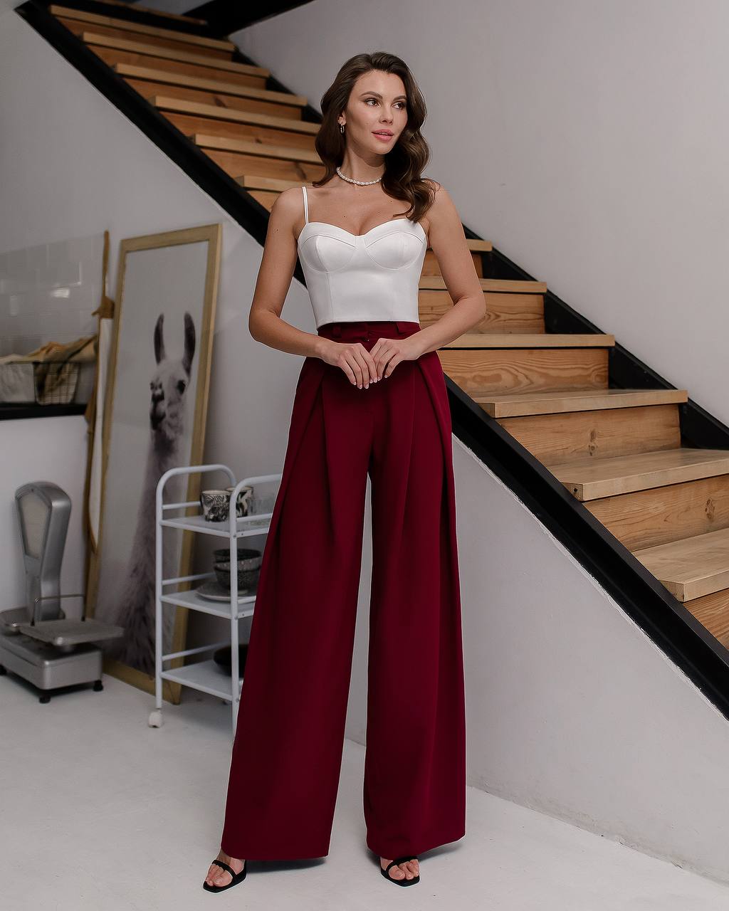 Woman in a white top and red pants standing in a modern interior setting with wooden stairs.