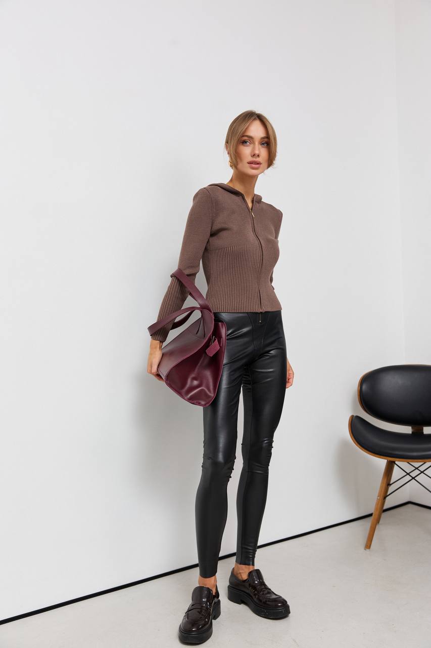 Woman in brown sweater and black leather pants holding a red bag against a white background