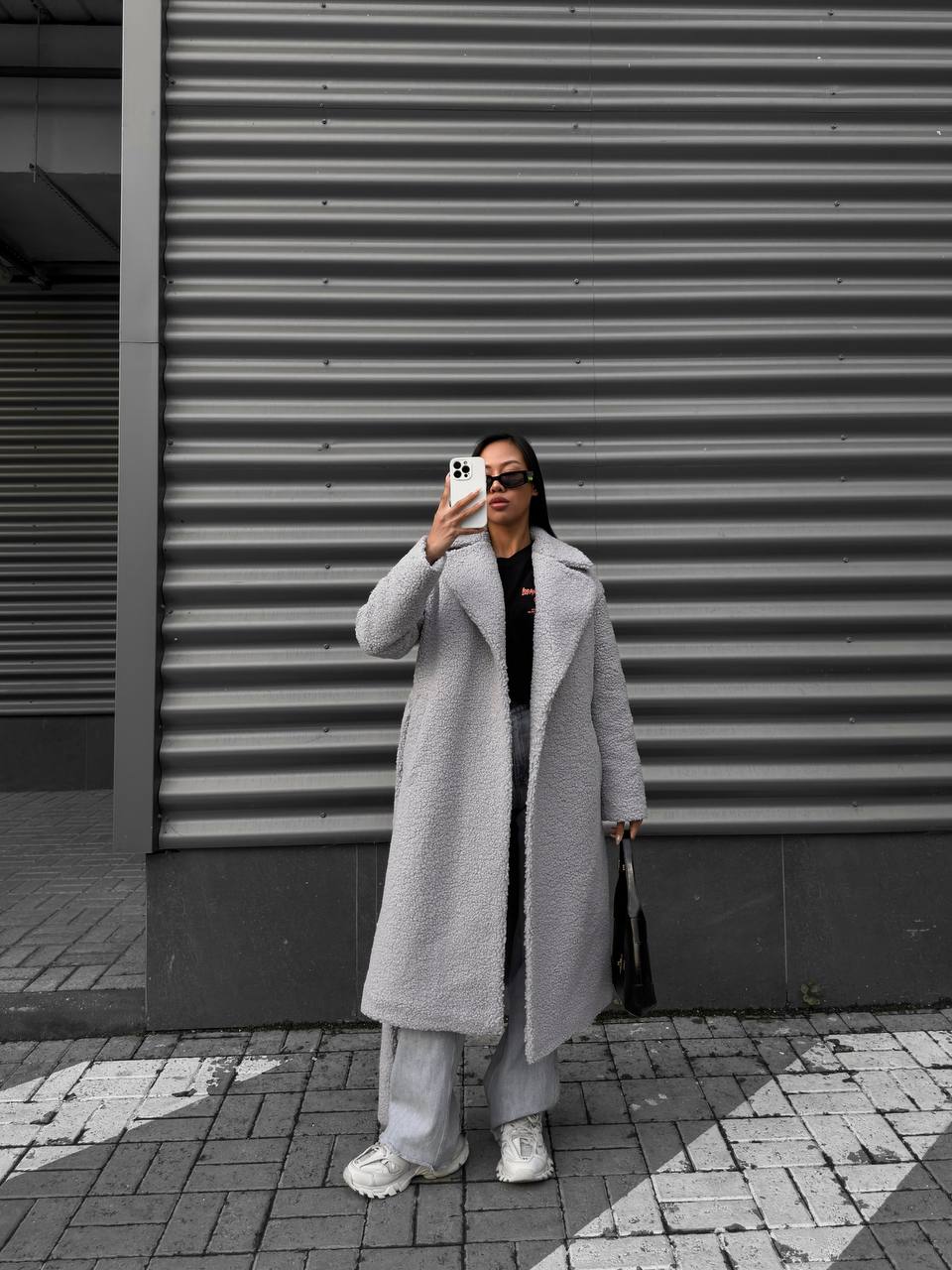 Person wearing a long gray coat standing in front of a corrugated metal wall.