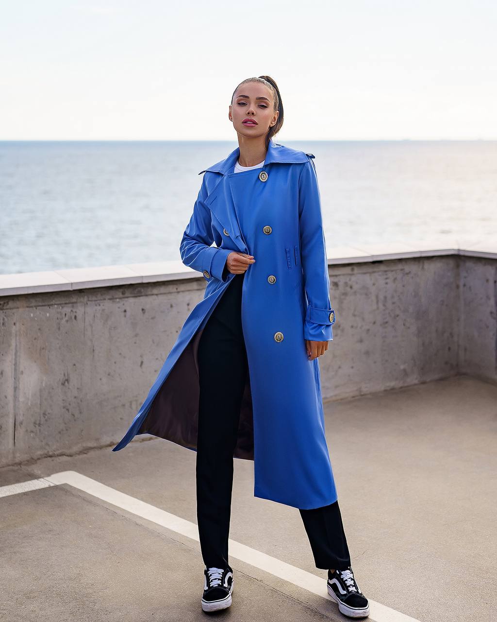 Person wearing a blue trench coat standing on a rooftop with a body of water in the background