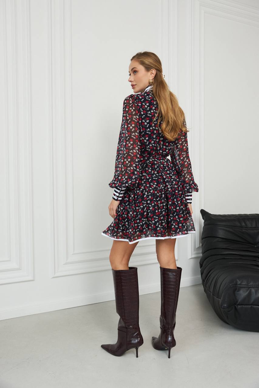 Woman in a patterned dress and knee-high boots standing in a room with a black chair.