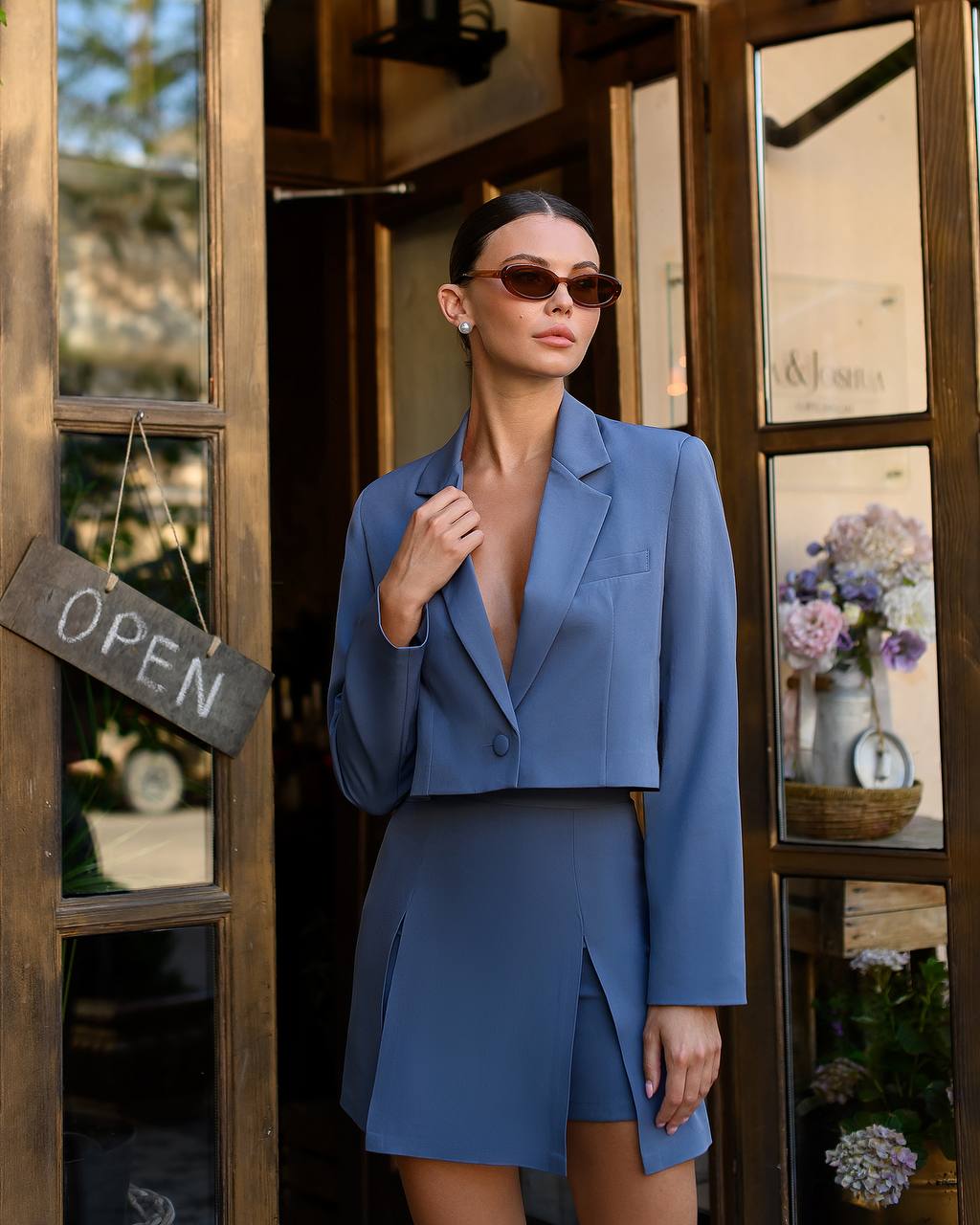 Woman in a blue outfit standing in front of a store entrance with 'OPEN' sign.