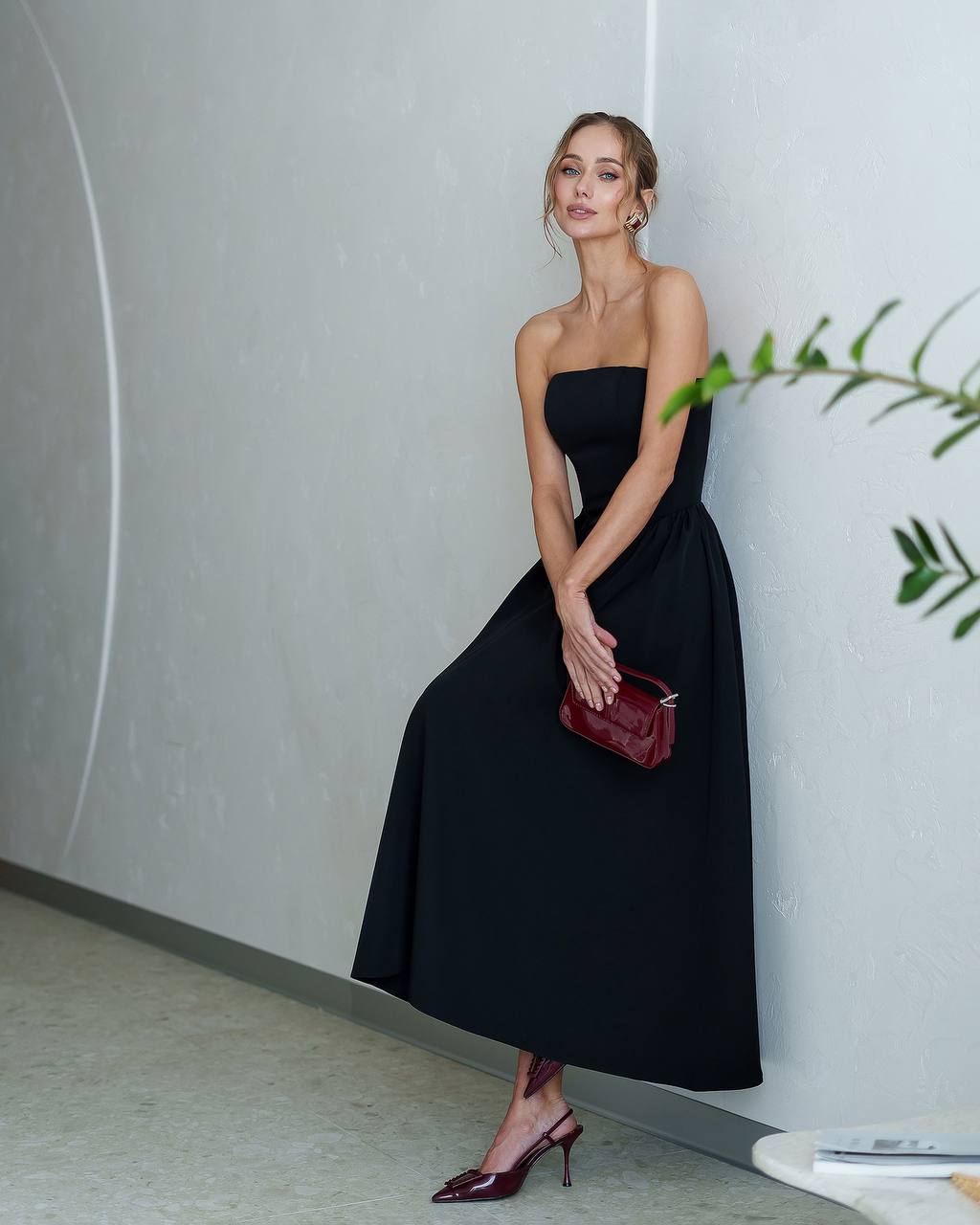 Woman in a black strapless dress holding a red clutch against a light gray wall.
