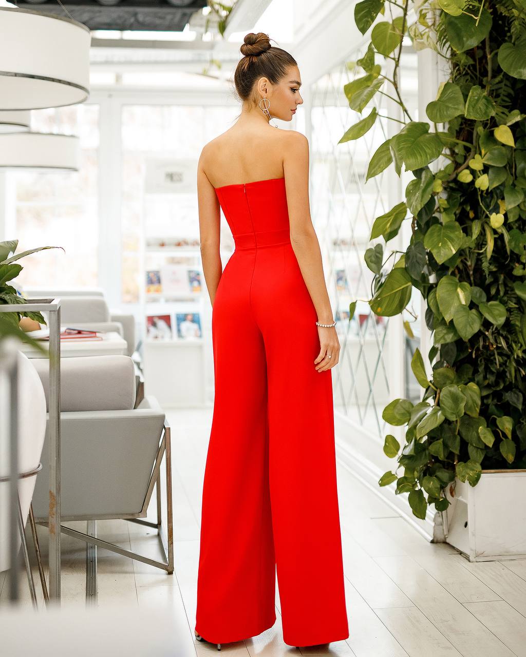 Woman in a red strapless jumpsuit standing in a modern indoor setting with plants.