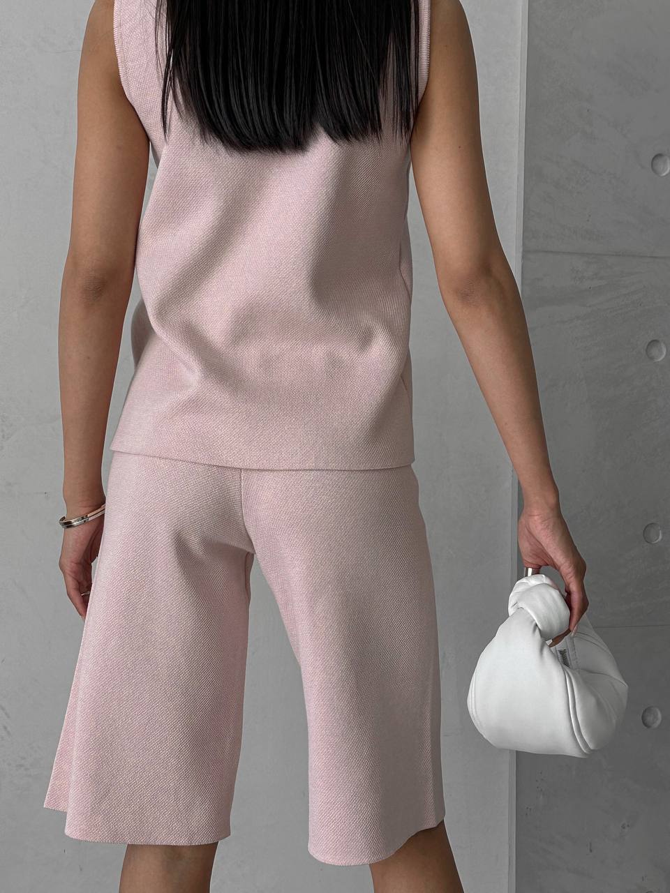 Person wearing a pink sleeveless top and shorts set, holding a white handbag against a gray background.