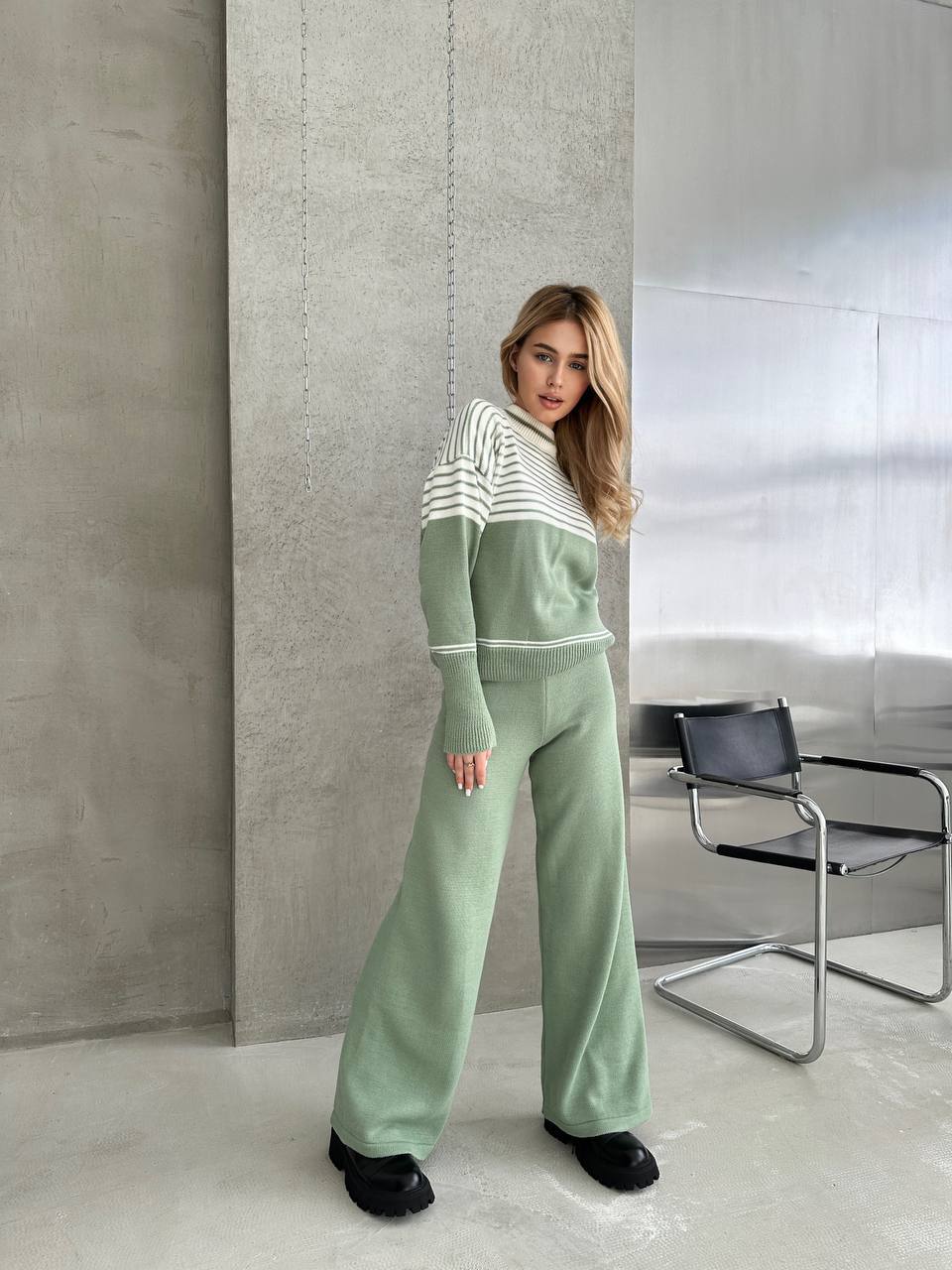 Woman wearing a green tracksuit in a modern room with a chair and concrete wall.