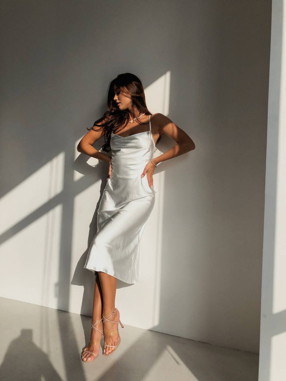 Woman in a light dress standing against a gray wall with sunlight casting shadows.