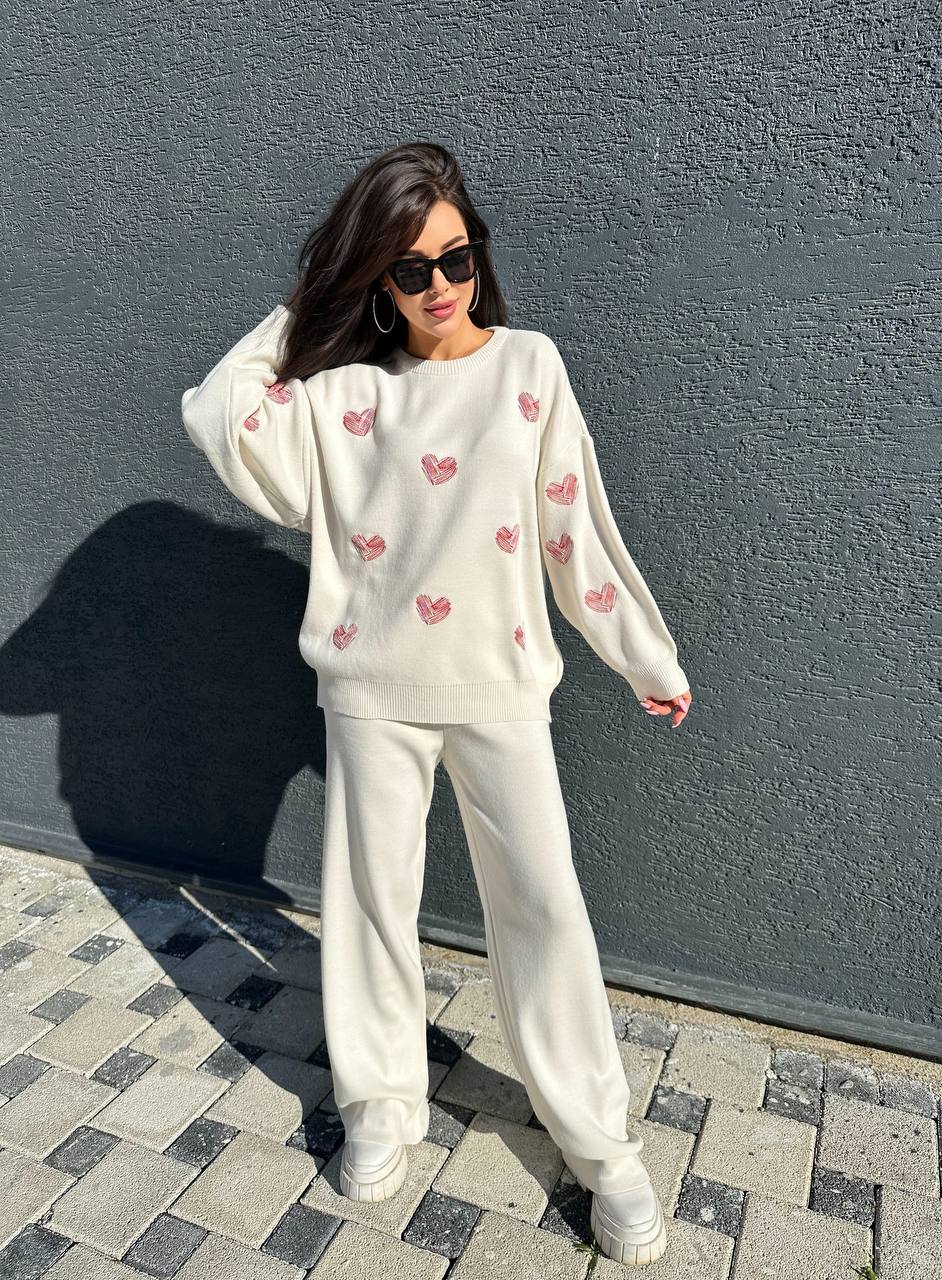 Woman wearing a white sweatshirt with pink heart patterns and white pants against a gray wall.