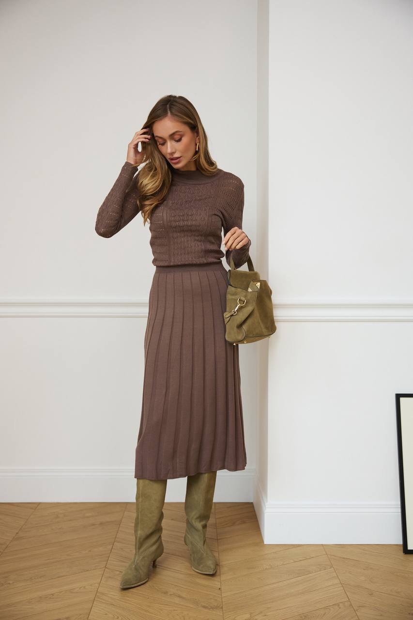 Woman in a brown ribbed top and skirt holding a green handbag against a white wall.