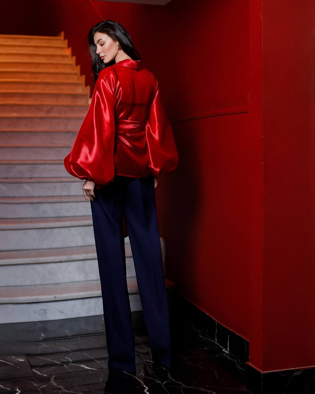 Woman in a red satin blouse and blue pants standing on a staircase with a red wall.