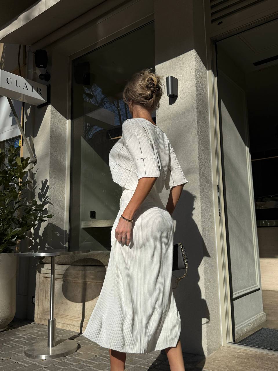 Woman in a white dress standing outside a building with a plant and table in the foreground.