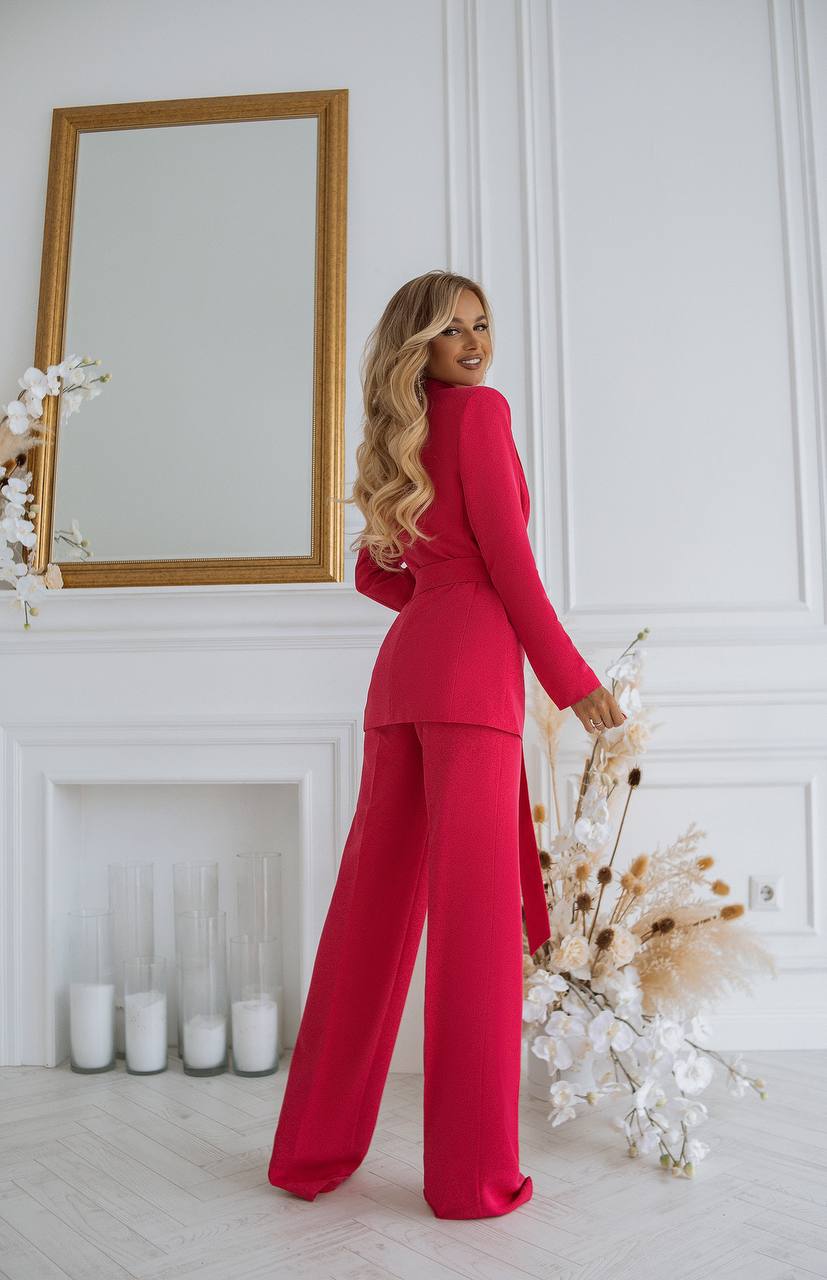 Woman in a red outfit standing in a stylish room with a mirror and decorative plants.