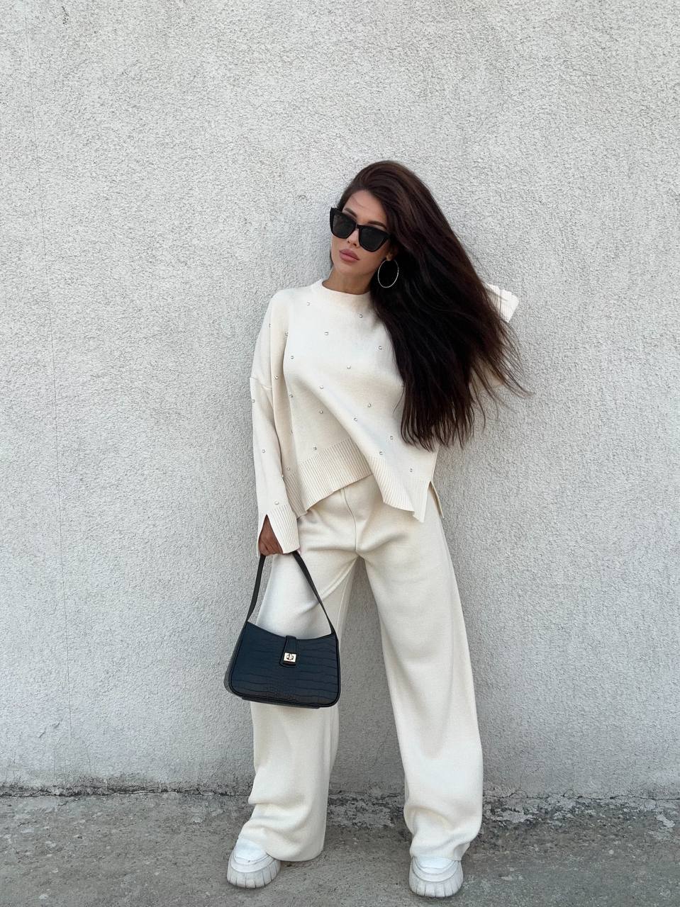 Person wearing a white outfit with a black handbag against a gray wall