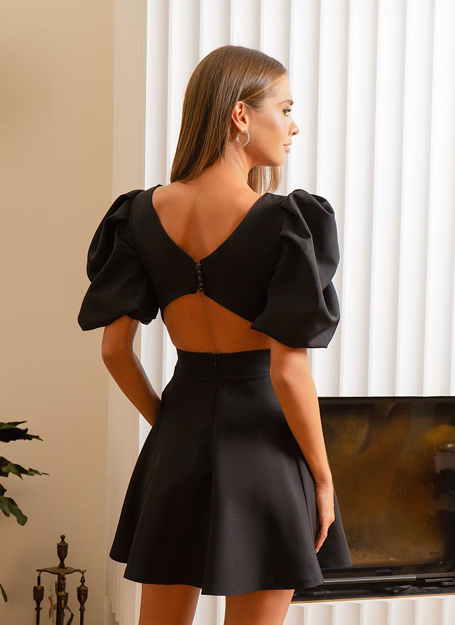 Woman wearing a black dress with puffed sleeves in a room with white curtains.