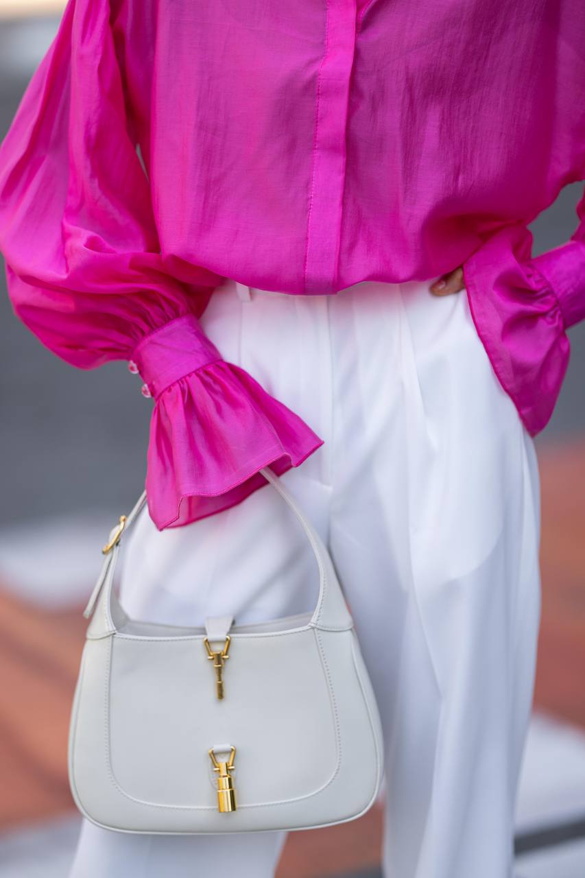Person wearing a bright pink blouse and white pants, holding a beige handbag.