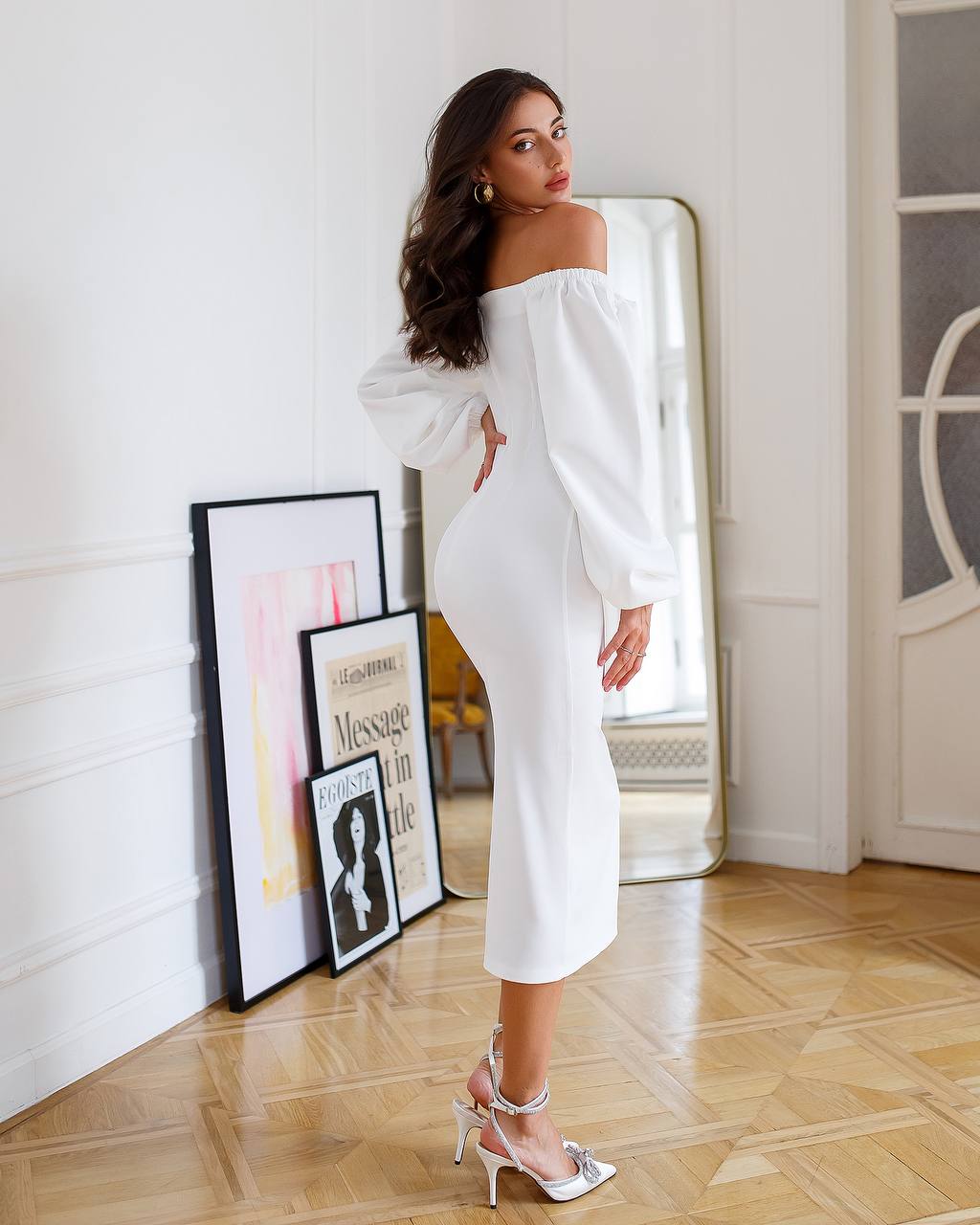 Woman in a white off-shoulder dress standing in a room with framed pictures on the wall.