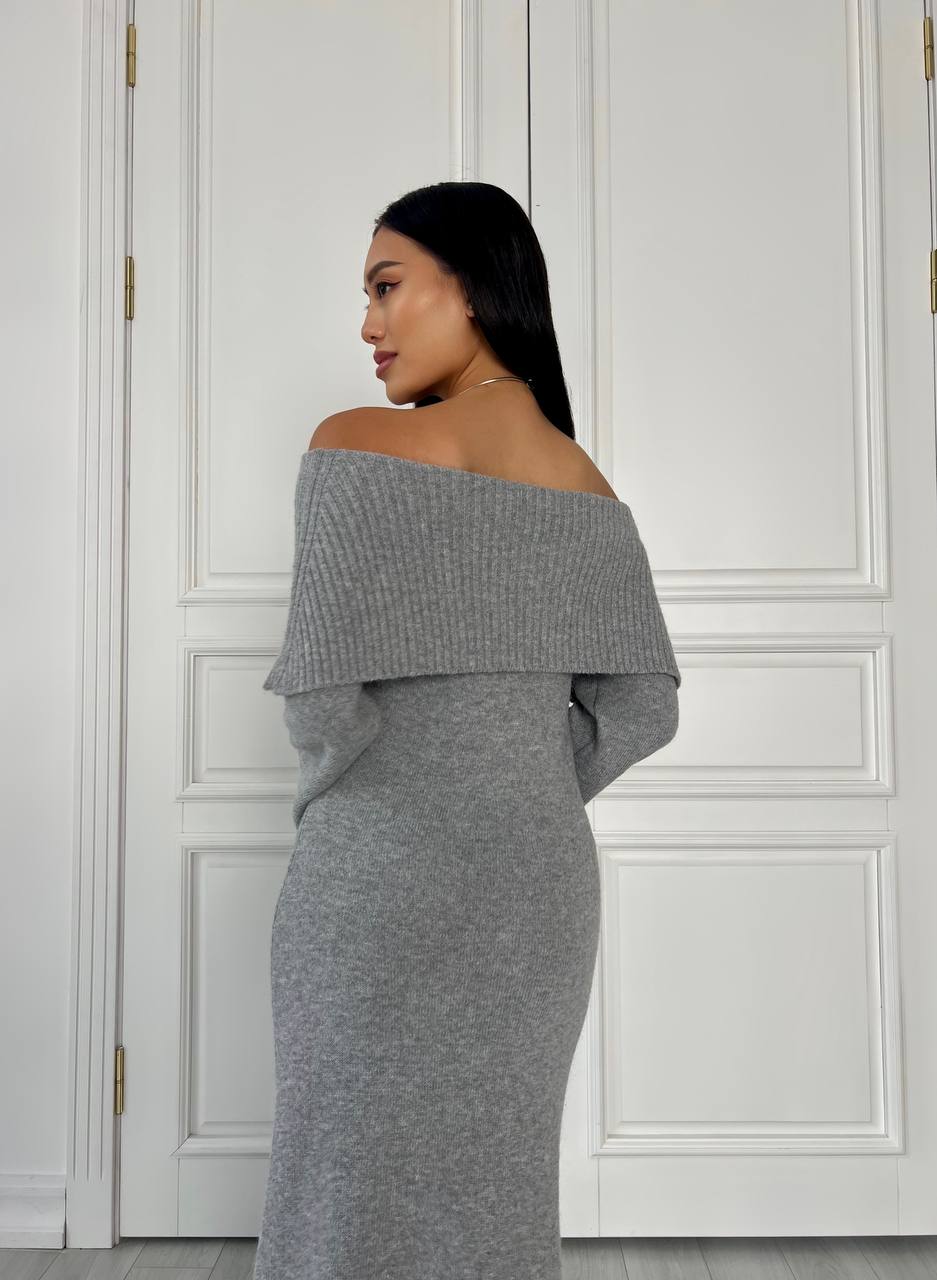Woman wearing a gray off-shoulder dress against a white paneled wall.