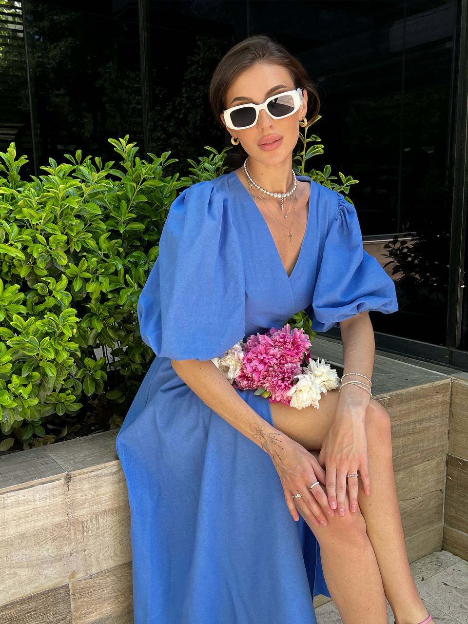 Woman in a blue dress sitting outdoors with flowers, wearing sunglasses.