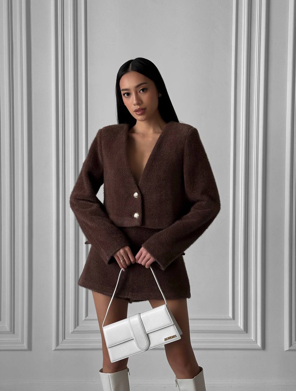 Woman in a brown outfit holding a white handbag against a gray paneled wall.