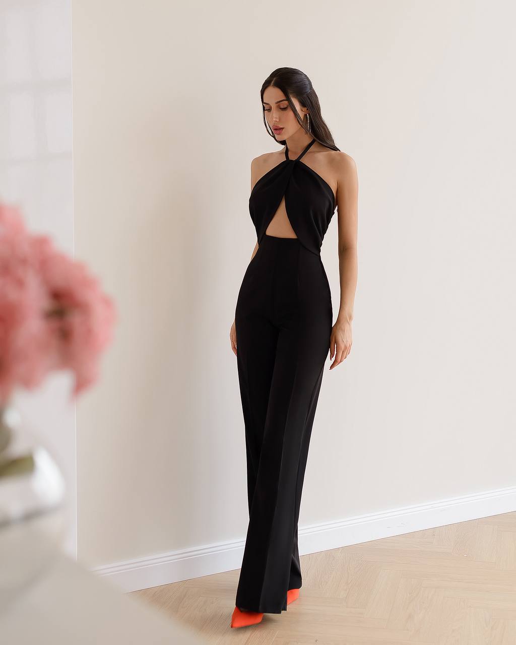 Woman wearing a black halter neck jumpsuit with cut-out details, standing against a plain wall.