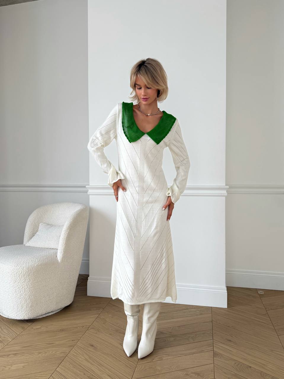 Woman wearing a white dress with a green collar in a room with a white wall and a white chair.