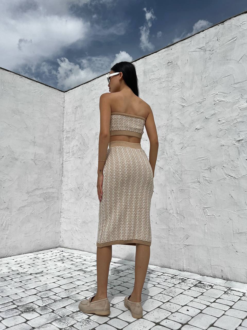 Woman in a patterned outfit standing in a white-walled room with a blue sky outside.