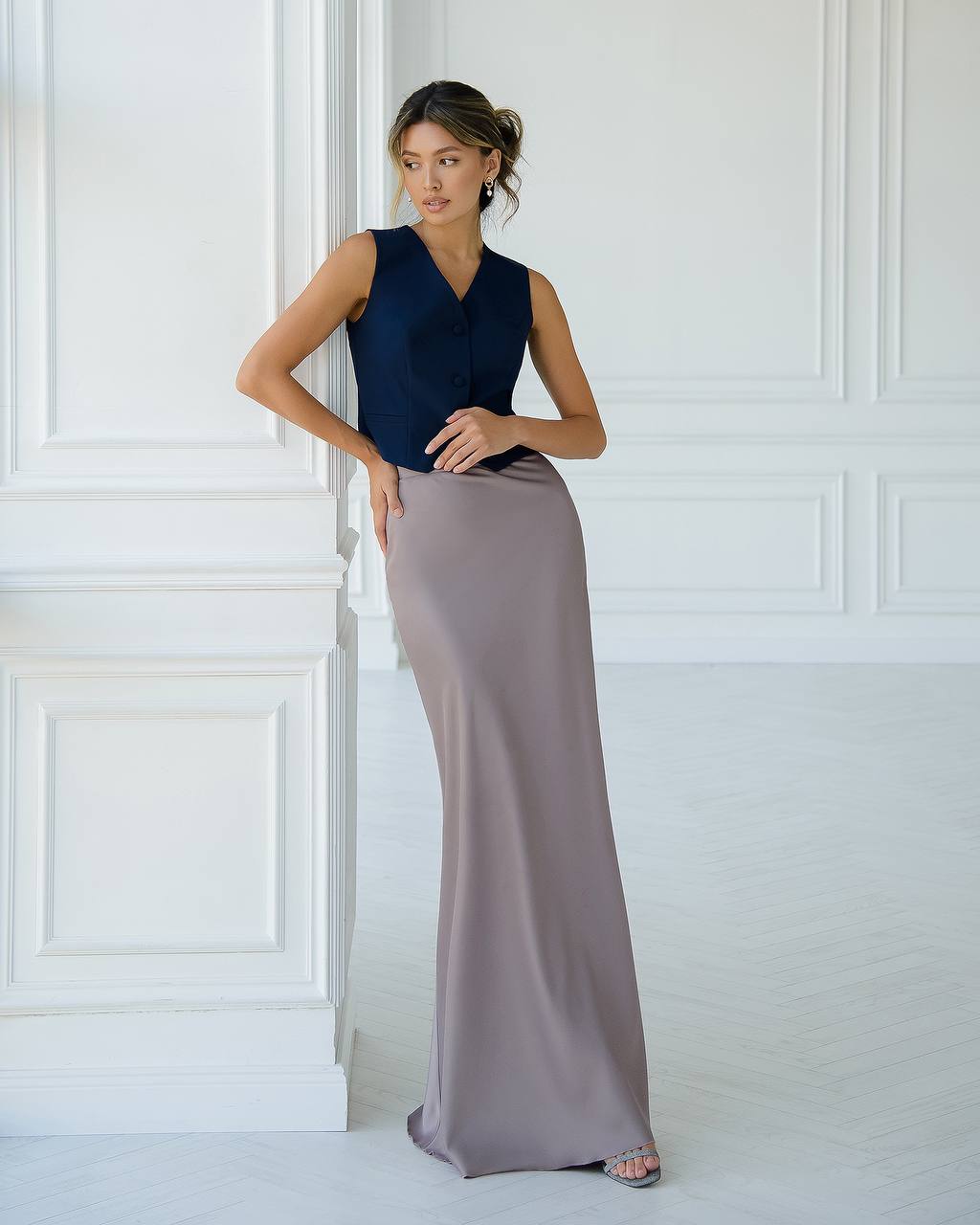 Woman wearing a navy sleeveless top and long beige skirt against a white paneled wall.
