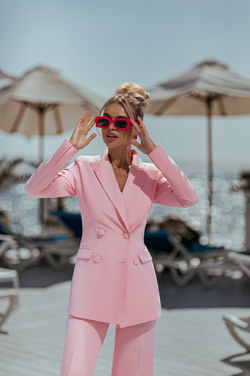 Woman in a pink suit and sunglasses on a beach with umbrellas and chairs in the background.