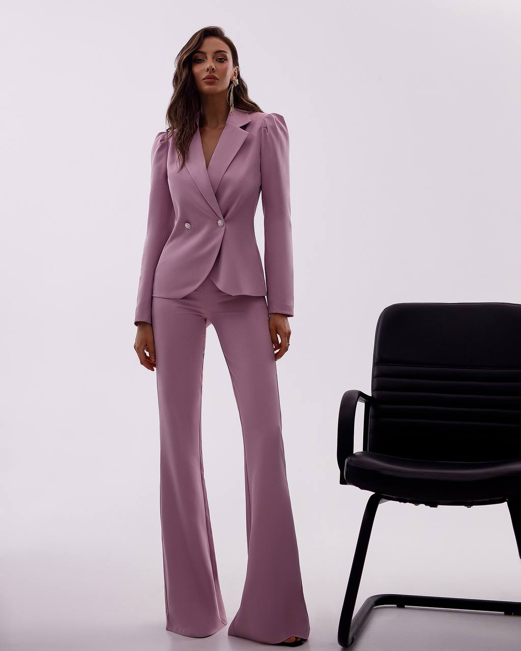Woman wearing a pink suit standing next to a black chair on a white background