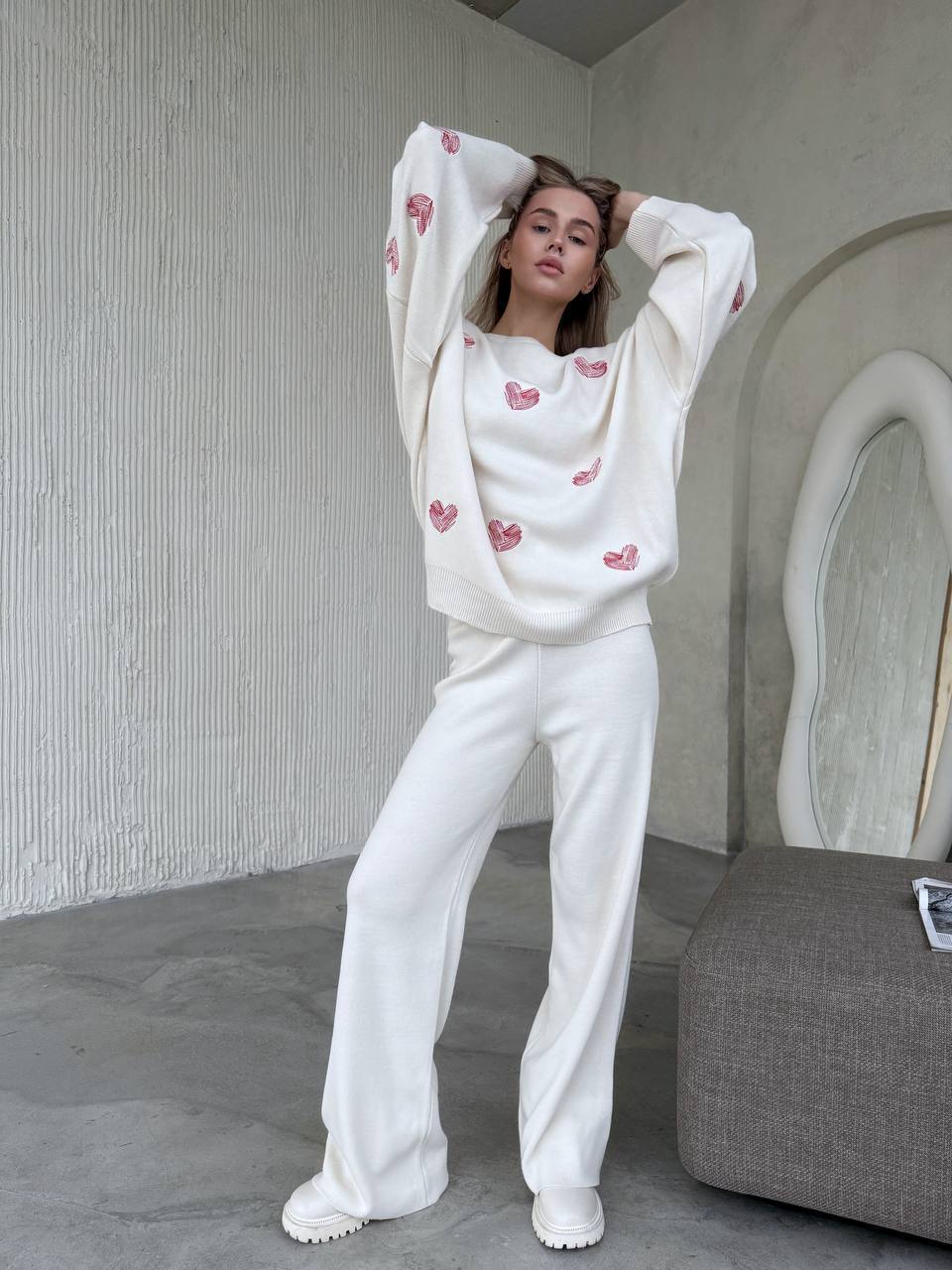 Person wearing a white sweatshirt with heart patterns and white pants in a minimalistic room.
