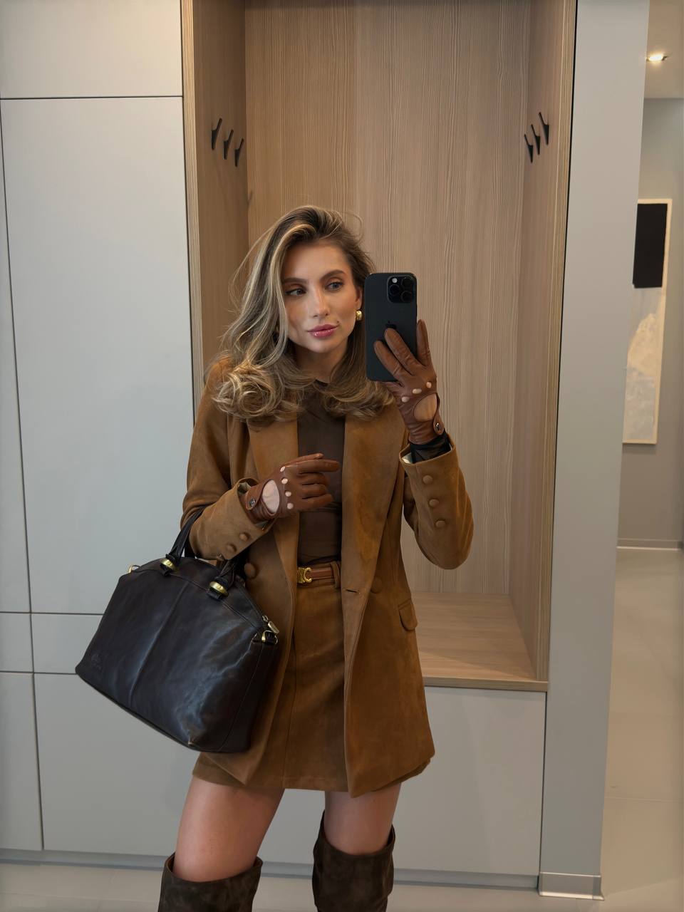 Woman taking a mirror selfie wearing a brown coat and holding a black handbag.