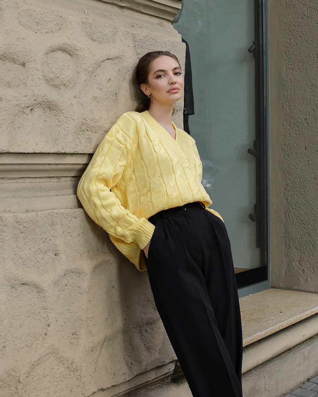 Woman wearing a yellow sweater and black pants leaning against a stone wall.