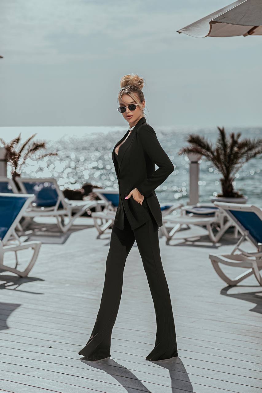 Woman in a black suit standing on a beach with lounge chairs and umbrellas in the background.