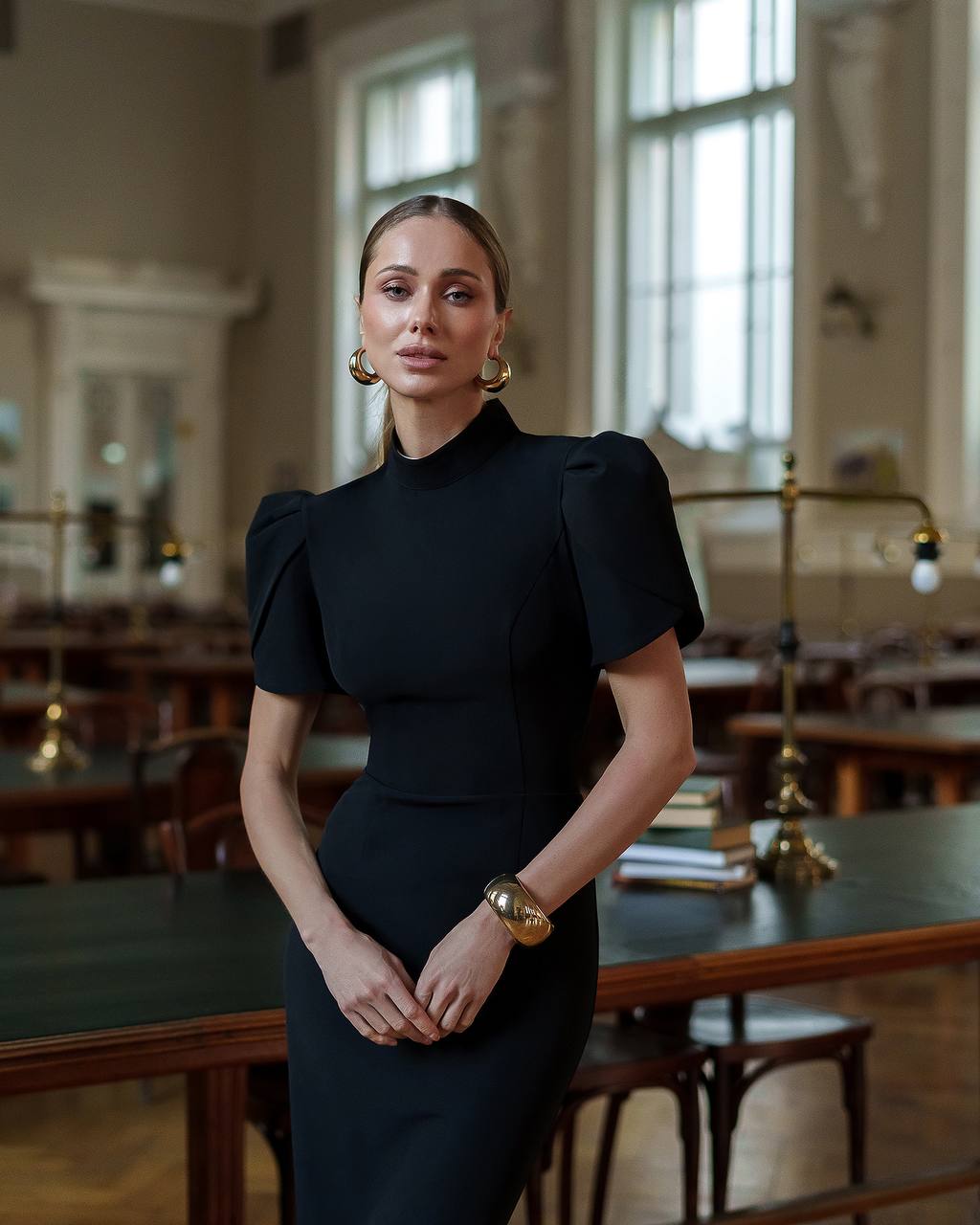 Woman in a black dress standing in a room with large windows and wooden tables.