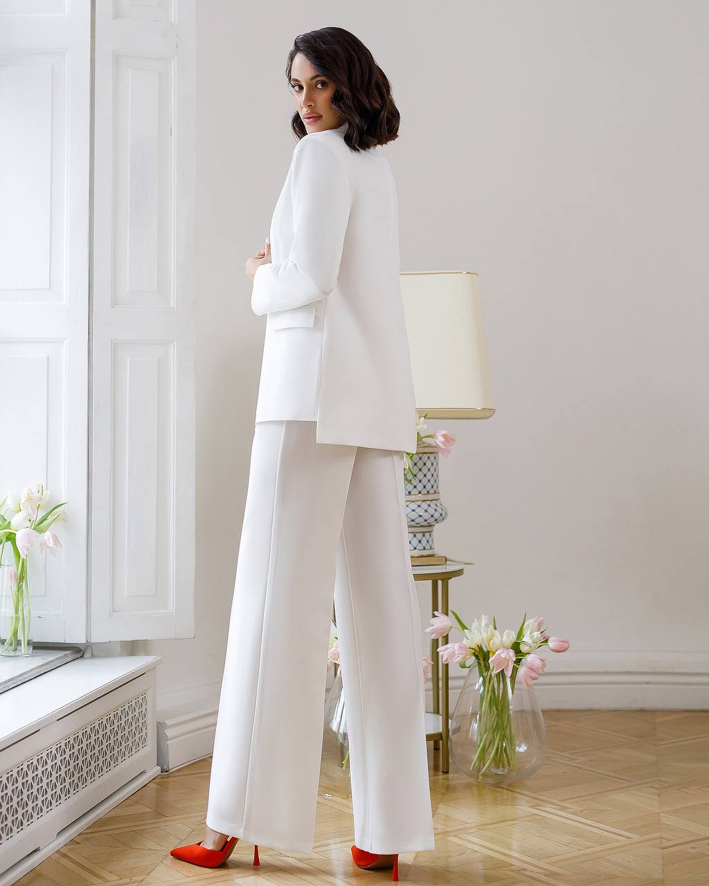 Woman in a white suit standing in a room with flowers and a lamp.