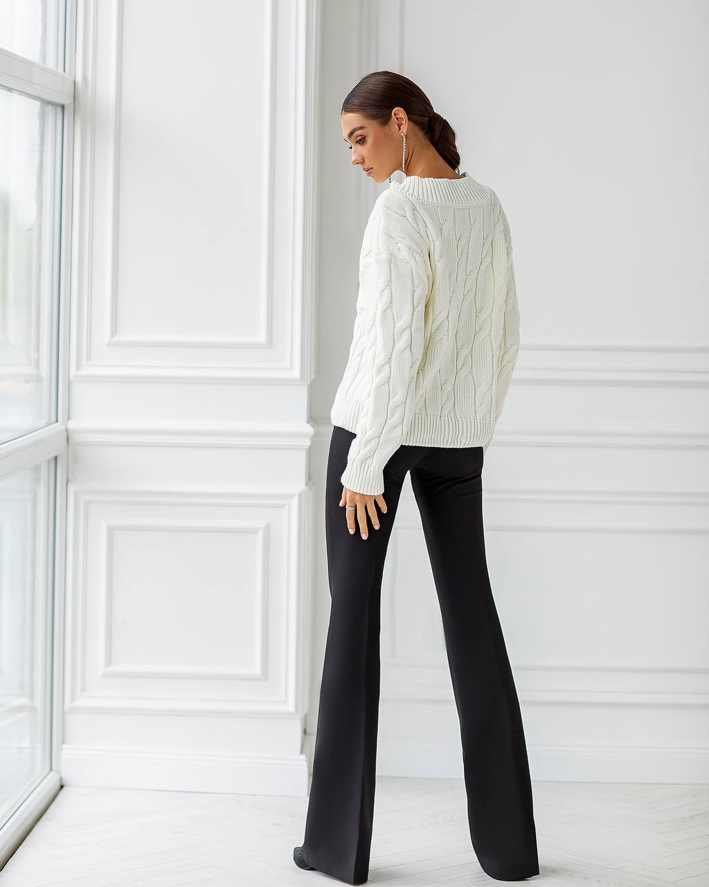 Woman wearing a white cable knit sweater and black pants in a minimalistic room.
