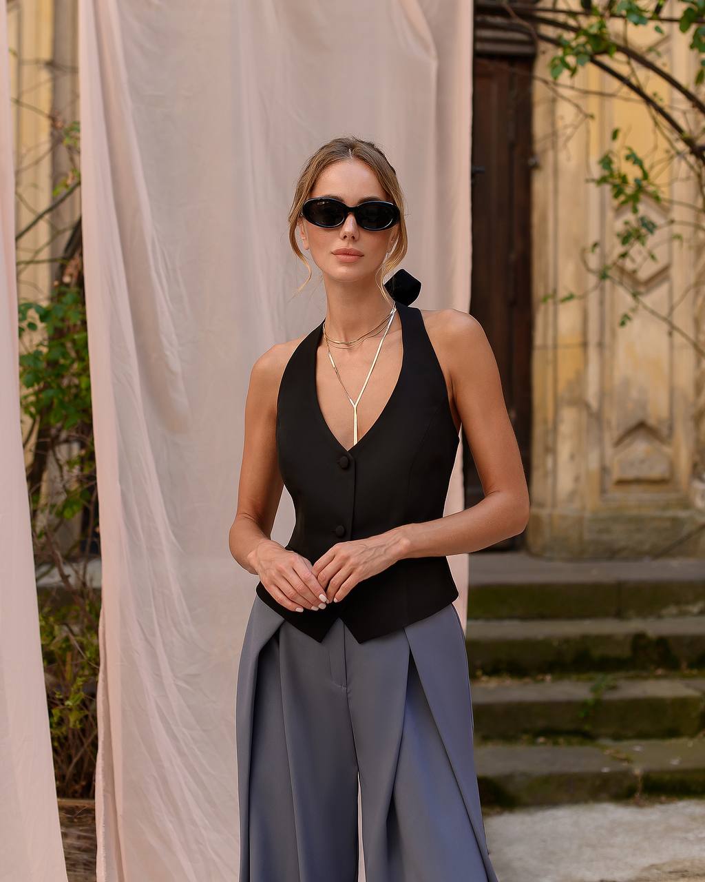 Woman wearing a black top and gray pants standing in front of a beige curtain.