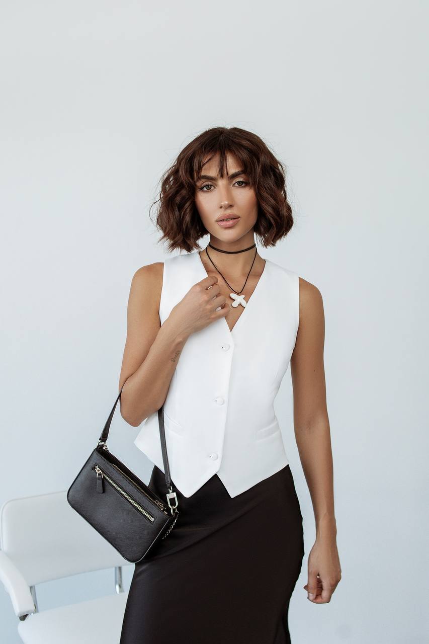 Woman wearing a white sleeveless blouse and black skirt, holding a black handbag against a light background