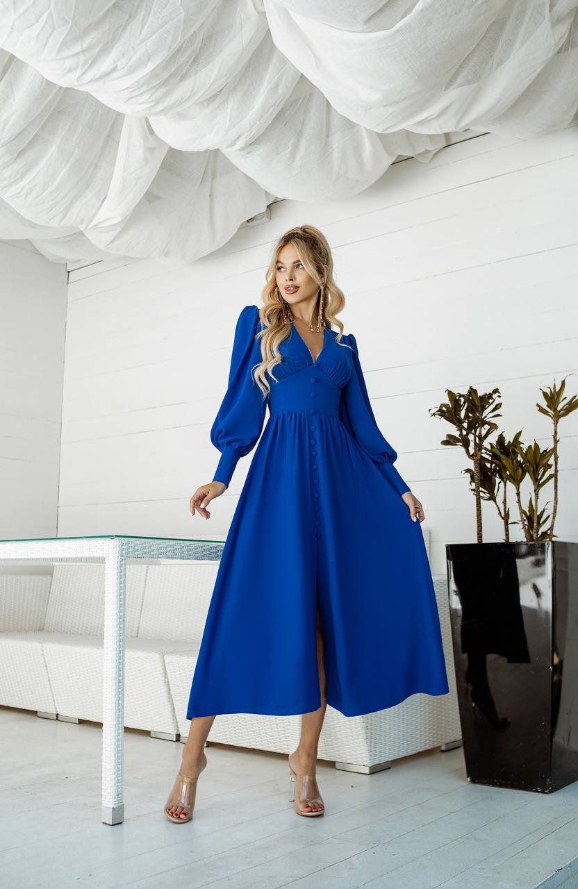 Woman in a blue dress standing in a modern indoor setting with white walls and a glass table.