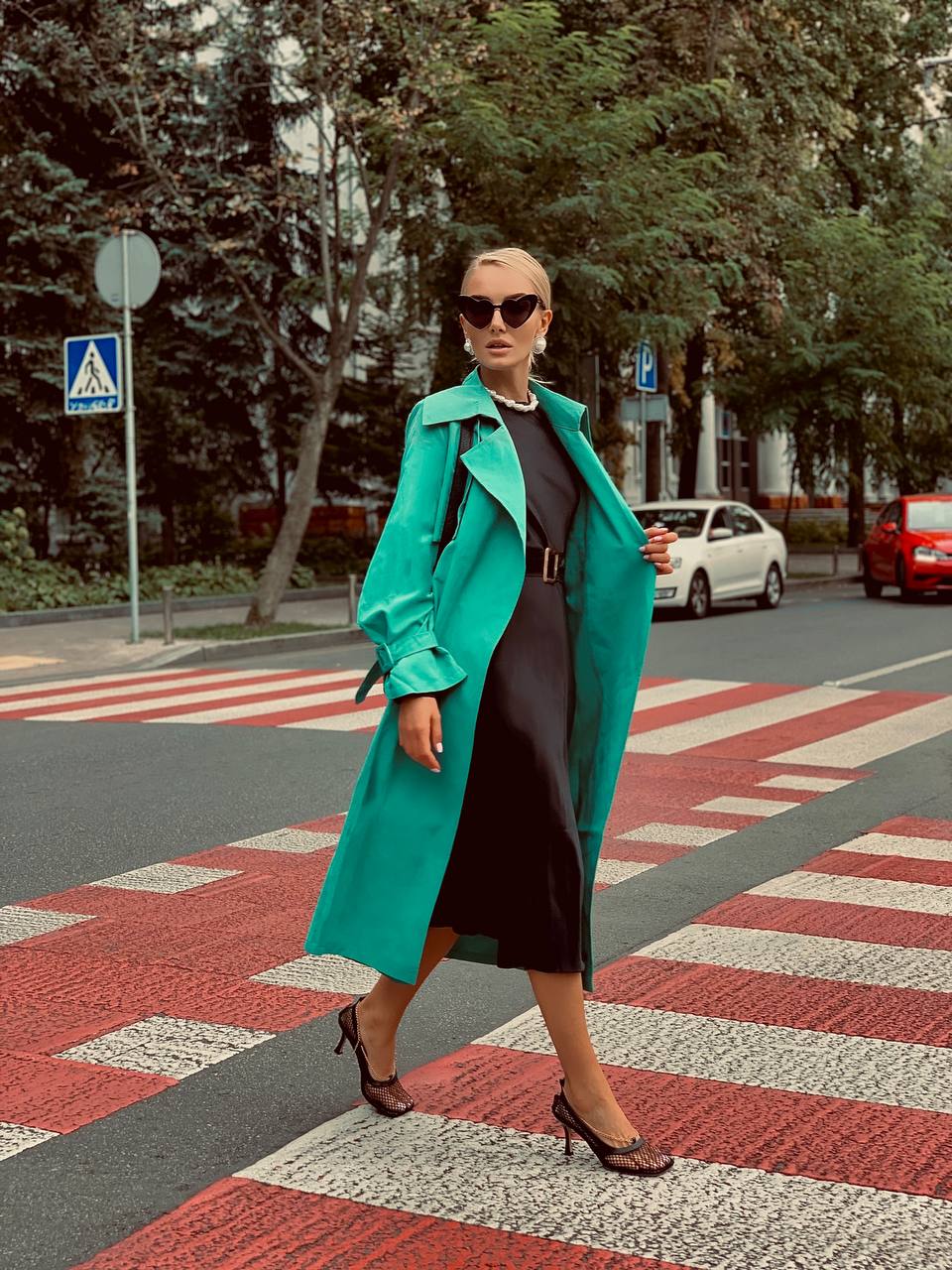 Woman in a teal coat crossing a street with trees and cars in the background
